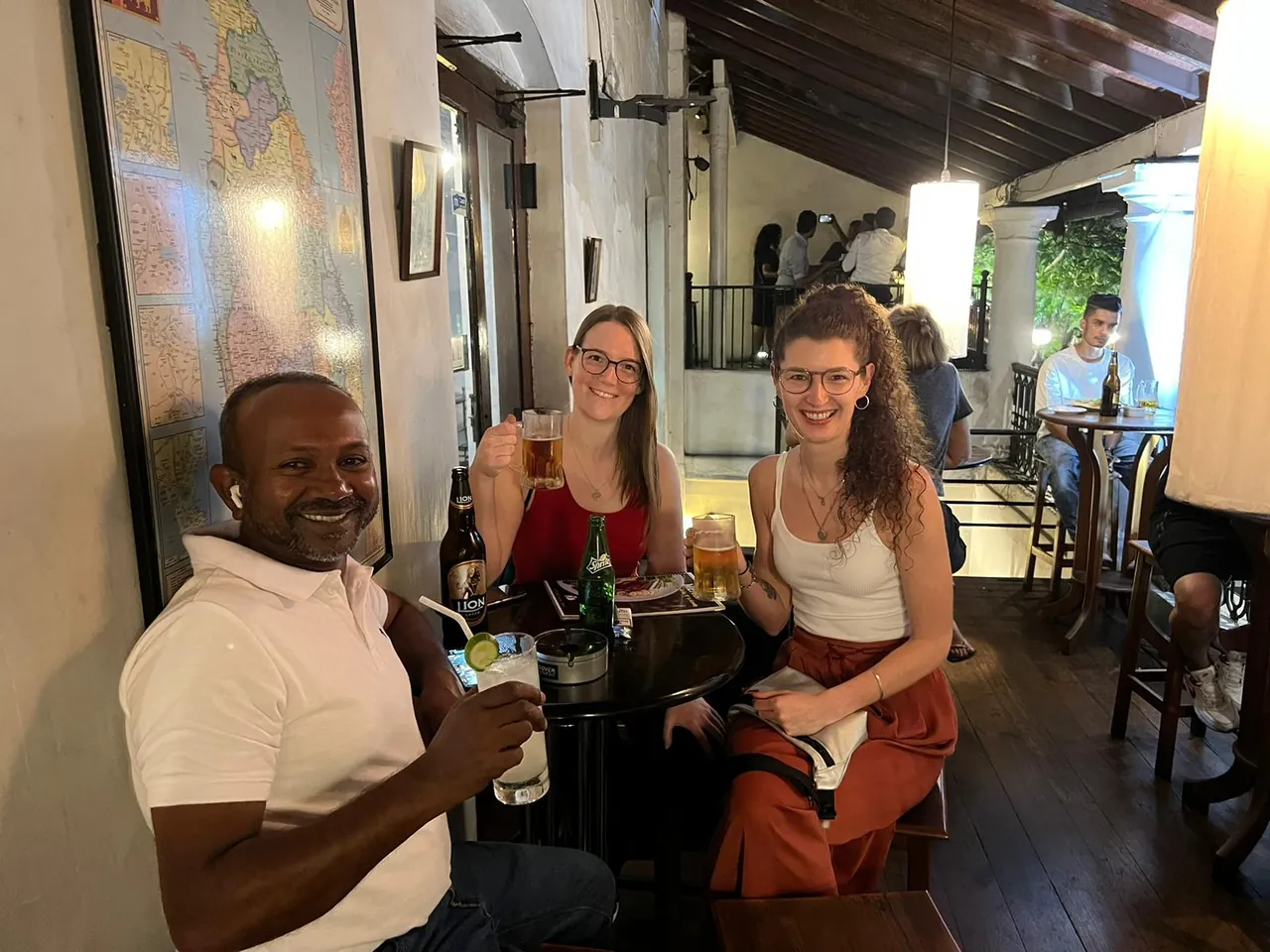 Reiseleiter Romesh stößt mit zwei weiblichen Reisegästen bei einem Bier in einer gemütlichen Bar in Sri Lanka an.